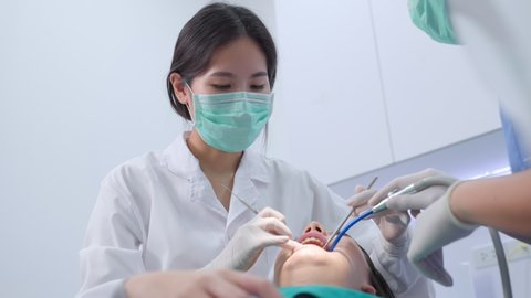 Asian young dentist wearing face mask, using medical instruments for oral care treatment. Assistant sitting beside, using suction machine. Medical service occupation and health care in dental clinic.