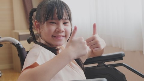 Portrait of Asian little illness girl sitting on wheelchair with smiling in hospital while waiting for medical service. The young kid feeling good, hopeful and thumbs up with cheerful and happy face.