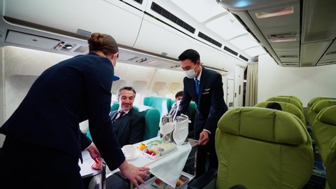 Asian male and female flight attendants in face masks for protective Covid-19 served airline meals to middle-aged Caucasian and Asian male passengers on seats. Some travelers don't wear a face mask.