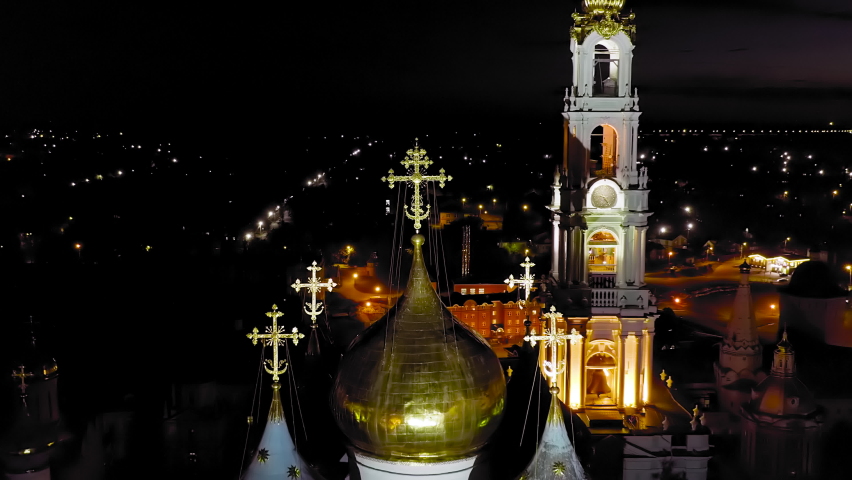 Sergiev Posad, Russia. The Trinity Lavra of St. Sergius rises above the city in a sweeping aerial night view, its illuminated five-tiered bell tower and gilded onion domes crowned with ornate Orthodox crosses standing out against the dark sky. Founded in 1337 by St. Sergius of Radonezh, the lavra is the largest active male monastery of the Russian Orthodox Church and a UNESCO World Heritage Site. The drone camera glides forward and upward past the gleaming golden domes, drawing closer to the richly detailed cross at the summit while the warm glow of monastery lighting contrasts with the scattered city lights below. The surrounding fortified walls and historic rooftops fade into shadow, emphasizing the vertical ascent toward the monastery