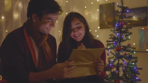 Shot of young attractive Indian Asian cheerful married couple is being excited after opening a Christmas present box in well-decorated interior house setup. festival holiday celebration concept. 