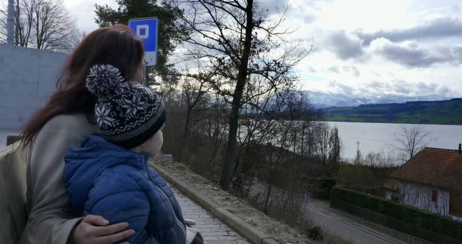 Mother and Son Sit Bench at Pull-in Parking near Highway Resting after Long Ride Watching at Picturesque Mountains and River.  Grey Overcast Sky. Eich Switzerland Europe