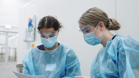 Handheld Medium Shot Of Two Young Female Doctors In Scrubs, Eyewear And Face Masks Checking Through Paperwork And Reaching For A File In Hospital