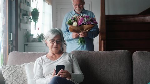 Old man giving flowers at his wife sitting on the sofa at home for the San Valentines’ day. Pensioners enjoying surprise together. In love people having fun.