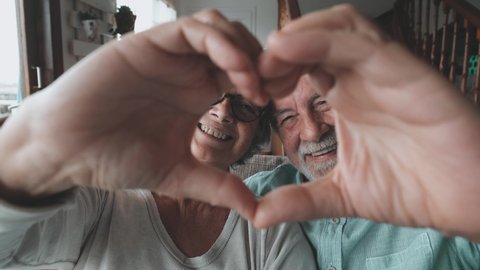Close up portrait happy sincere middle aged elderly retired family couple making heart gesture with fingers, showing love or demonstrating sincere feelings together indoors, looking at camera.
