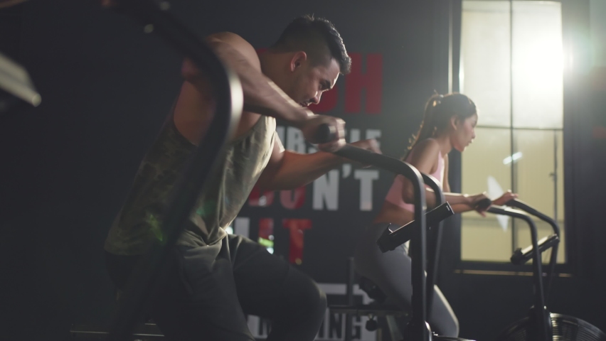 Close-up shot of people sitting on stationary bicycles or exercise bikes in stadium. Asian muscle man and woman spinning on cardio machine together. Health care and fitness in gym concept. Royalty-Free Stock Footage #1068628859