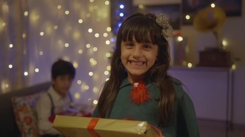Close shot of a cute little dressed up cheerful Indian girl holding a wrapped up present box in hand, smiling and looking at the camera on the occasion of Christmas eve in well decorated house setup