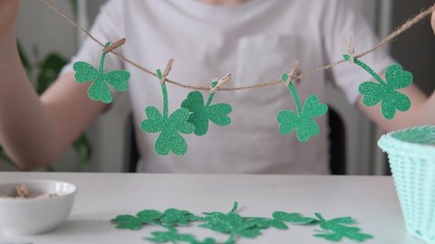 DIY St.Patricks Day decor. Hands hold shamrocks garland of glitter paper on white table. Slow motion
