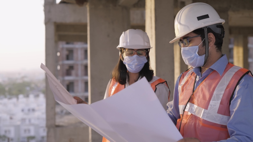 A young male and female civil engineers wearing protective face masks and safety helmets standing on an under-construction high rise building and discussing over work together amid COVID 19 pandemic. | Shutterstock HD Video #1068636455