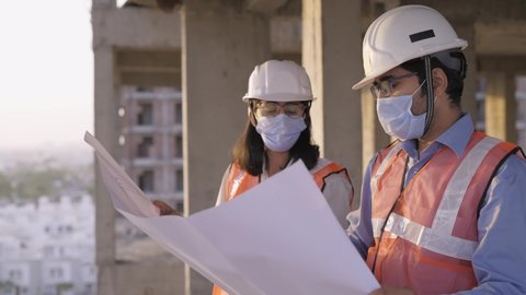 A young male and female civil engineers wearing protective face masks and safety helmets standing on an under-construction high rise building and discussing over work together amid COVID 19 pandemic.