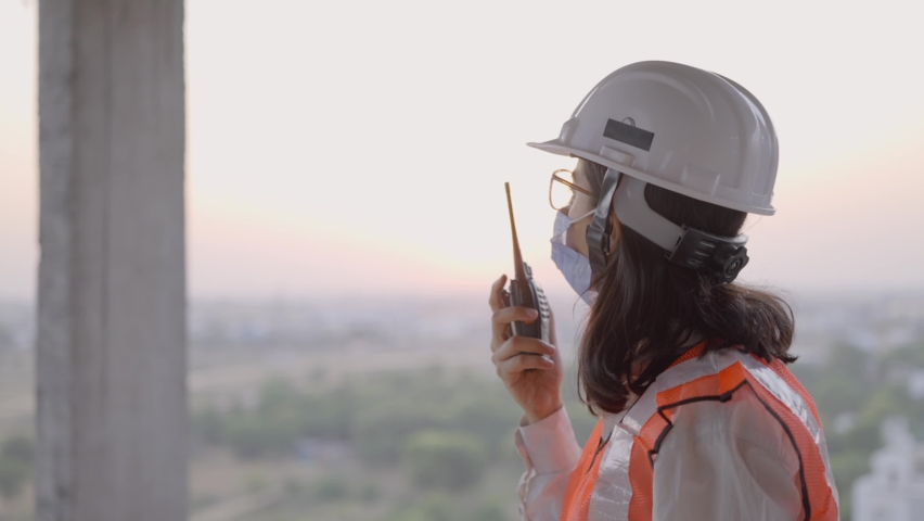 Close up shot of a young attractive Indian female civil engineer standing at the site wearing a protective face mask and a safety helmet supervising the construction activity using a walkie talkie  Royalty-Free Stock Footage #1068636458