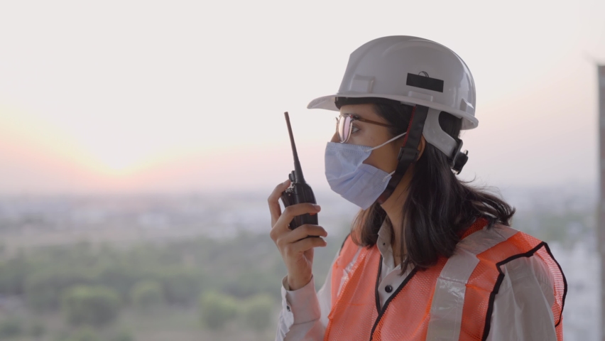Close up shot of a young attractive Indian female civil engineer standing at the site wearing a protective face mask and a safety helmet supervising the construction activity using a walkie talkie  | Shutterstock HD Video #1068636458