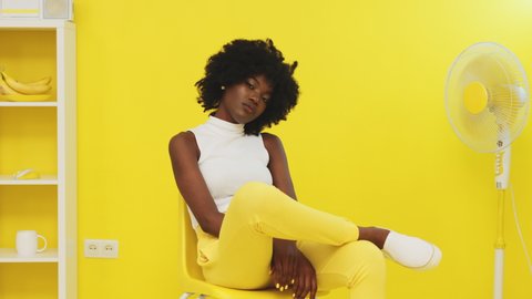 Portrait of black woman, young and beautiful model, sitting on yellow chair next to a fan, chilling out and looking at camera confidently, smiling and sharing positive vibes, Slow motion.