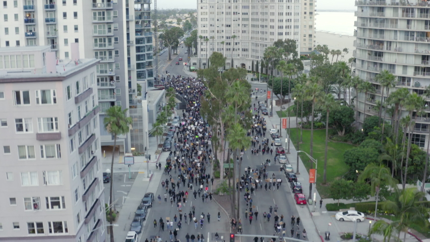 Rising Aerial: Huge Crowd Of Protesters Meets At City Crossroads With Signs Under Tall Apartment Buildings Near Beach