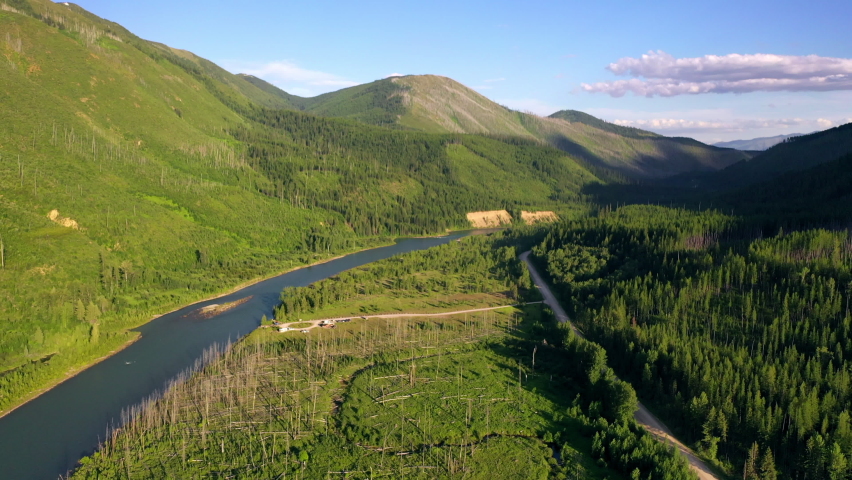 Aerial tilt down shot of river flowing through green forest against sky, drone flying forward towards mountains on sunny day - Great Northern Flats, MT
