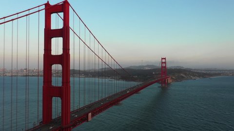 Aerial panning shot of vehicles on Golden Gate Bridge over bay, drone flying forward over famous landmark against sky at sunset - San Francisco, California