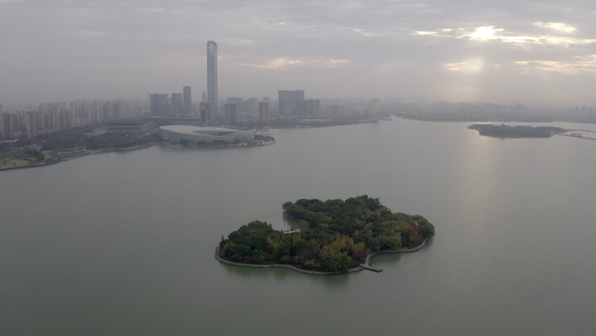 Aerial panning shot of island on Jinji Lake near city against cloudy sky, drone flying near modern buildings - Suzhou, China