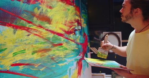 Close up of Caucasian young talented excited male contemporary painter working in studio emotionally drawing on large canvas using paint brush, abstract colorful artwork, artist concept