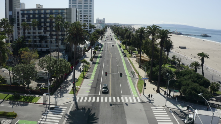 Aerial Forward: Traffic Moves Down A Long Beach Side Road Under Palm Trees In The Sunshine Near City