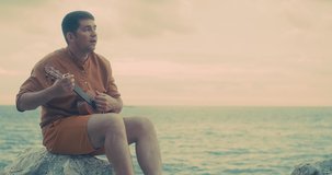 Fun attractive man traveler sitting on sea beach with ukulele under sunset sky in evening time. Male playing, sing music, relaxing enjoying holidays, travel day. 4K video
