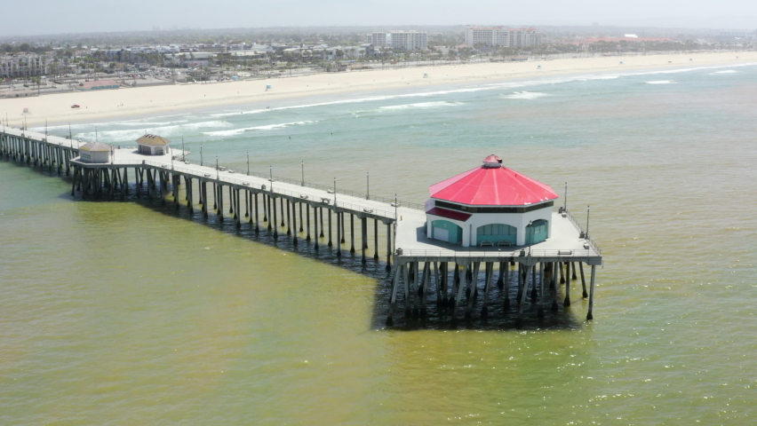 Aerial Pan: Long Pier Stretches Into Green Waters Of Pacific Ocean On Sunny Day With Distant Beach And Cityscape