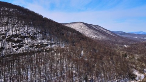 Aerial drone footage of a snowy mountain valley in early spring on a sunny day in the Appalachian Mountain Range, just after winter ends with forests and snow and sunshine and blue skies