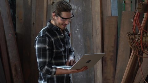 young hipster carpenter man holding computer laptop looking and checking wood in workshop . craftsman wearing safety glasses choosing plank