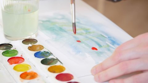 An amateur and self taught artist is painting an abstract watercolor picture on a white paper, washing out his brush in a glass of water and greasing it in a set of coulors and smearing the sheet
