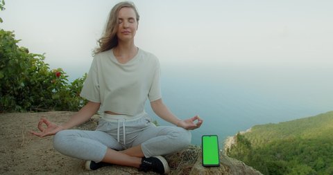 Woman relaxed meditate in yoga pose with green screen smartphone and sea background