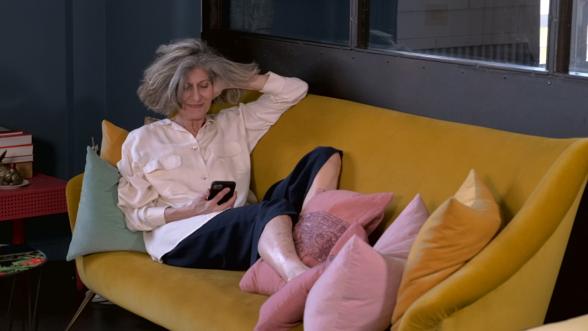 Senior womaan relxing on sofa, reading on her smart phone