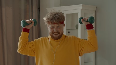 CARSH ZOOM Portrait of funny overweight Caucasian man working out with dumbbells at home, looking into camera. Bright, commercial high key advertising style lighting