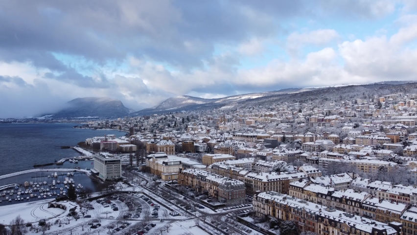 Drone fly over footage of Neuchatel city in Switzerland covered by snow in winter, with blue sky and clouds, cityscape, lake view and Jura mountains in the distance