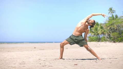 Young attractive millennial man practicing yoga stretching on the beach. Concept of wellbeing, healthy, mindfulness wellness lifestyle.  Steady cam slow motion.