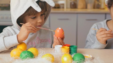 Happy Easter, kids. Girl and boy with rabbit ears on their head paint eggs in the kitchen of the house. Children brother and sister have fun on the holiday
