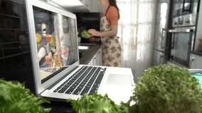 A young woman watching a online stream broadcast video blog tutorial about tasty and healthy food using laptop and cooking a meal at the home kitchen