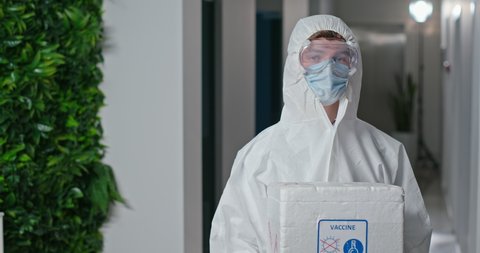 A young doctor in a white protective suit holds a thermo box with a refrigerator to transport ampoules with coronavirus vaccine