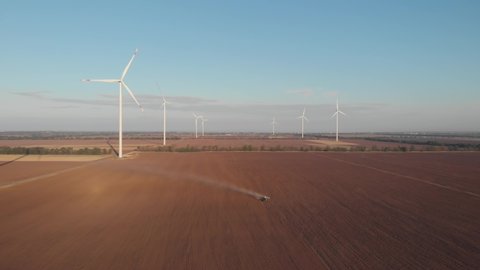 Harvester tractor plows agricultural field at wind turbines power plant farm. Wind mills rotating generating renewable energy at sunrise as tractor plows. Cinematic rural green energy 4K