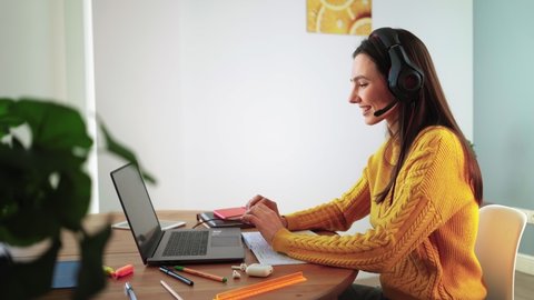 Happy woman in headphones is engaged e learning by web cam chat at home. Young female student in yellow sweater on laptop communicate online by video call. Distance education and Modern tech concept.