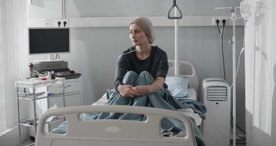 Young woman with cancer sitting alone on hospital bed during chemotherapy. Portrait of sick female patient with headscarf and nasal oxygen tube resting in hospital ward | Shutterstock HD Video #1068681881
