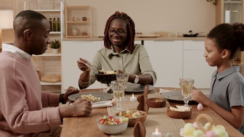 Chest-up of young happy African woman smiling, holding bowl with salad, filling plates of daughter and husband sitting at table on her sides. Family having festive dinner