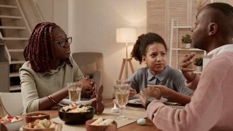 Medium close-up of school-aged African girl wearing hair up in ponytail, sitting at dinner table on festive night, looking at and listening to young mother and father having lively conversation
