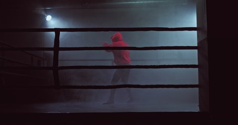 Male fighter is training in the ring, practicing punches, tubers of smoke. Fighter in a hood on a trainer, shadow boxing. 4k, ProRes