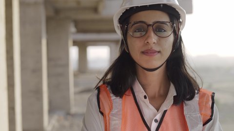 Track in a close-up shot of a young confident Indian Asian female civil engineer wearing a safety helmet standing with folded arms and looking at the camera on top of an under-construction building.