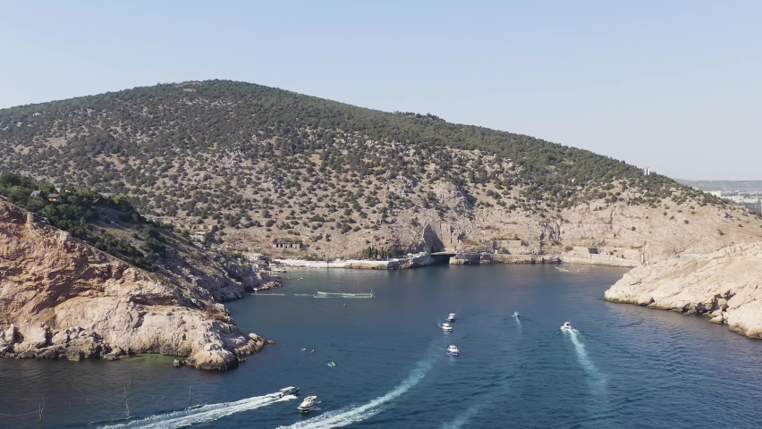 Sevastopol, Crimea. Balaklava Bay entrance features a massive concrete submarine tunnel carved directly into the rocky hillside, forming the visible mouth of a former Cold War underground naval complex. The camera glides over deep blue water where yachts and pleasure boats navigate the narrow inlet, leaving white wakes across the surface. Steep, arid hillsides with sparse vegetation rise on both sides of the bay, their rocky faces showing layers of geological strata. A winding coastal road traces the shoreline, with small structures and a memorial visible along the embankment. The scene transitions smoothly from the fortified tunnel entrance toward the inner harbor, revealing a compact town with white and pastel buildings clustered along the waterfront, red rooftops catching the sunlight, and a marina filled with docked vessels. The bright summer sky and clear atmospheric conditions emphasize the contrast between the deep turquoise water, tan rocky cliffs, and the built environment. The aerial perspective captures the strategic geography that made this location a top-secret military facility during the Soviet era, now repurposed as a naval museum complex.
