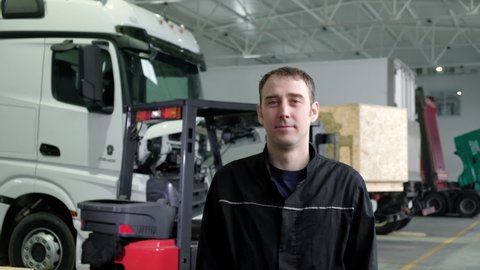 Portrait of mechanical engineer in uniform standing in a repair shop against the background of trucks looks at the camera and smiles. The loader operator works in the logistics of an auto repair shop