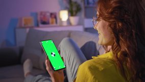 green vertical chromakey on smartphone, charming girl uses an Internet browser to watch video or an online store at home on couch while relaxing in evening