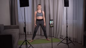 A young woman sits at home on a rug and teaches yoga online. Broadcast by phone. LED panels and a smartphone on a tripod. Video studio concept for vlogging.