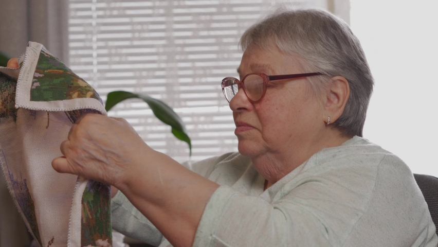 An elderly woman in glasses looking at his embroidery needlework. Hobby time at home concept