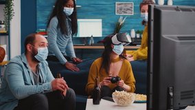 Woman playing video games using virtual reality headset wearing face mask during social pandemic to prevent illness with covid spending time with friends drinking beer. Multiethnic people hanging out