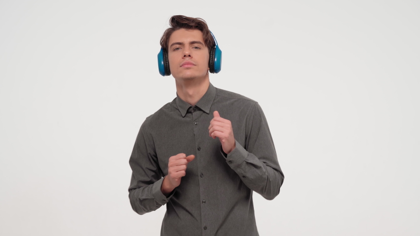 Shy dancer young man listening music wearing blue wireless headphones dressed in grey sports outfit. Funny dancing young guy isolated on white background. 4K footage. 